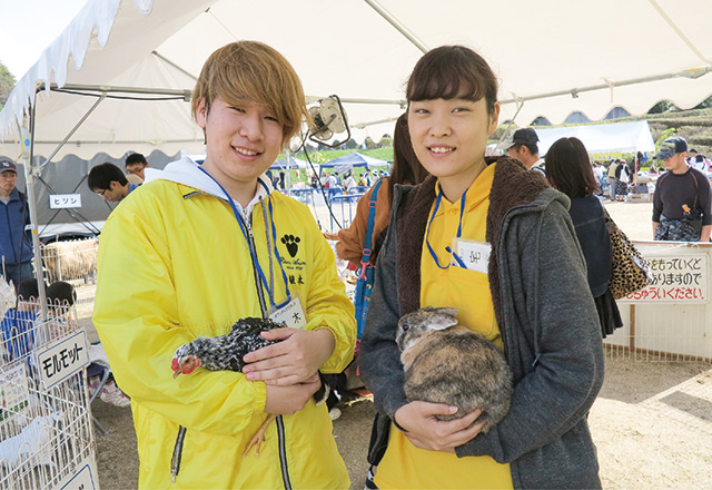 写真：動物愛護フェスティバルの様子。犬のきぐるみの横で笑顔でピースをしている学生。